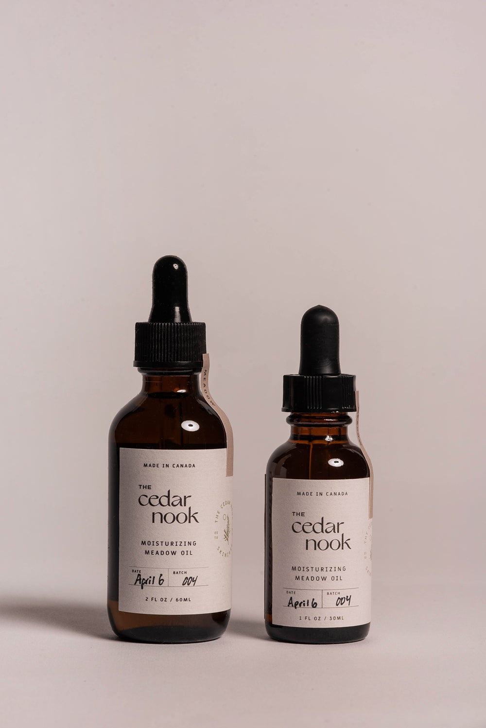 The Cedar Nook Moisturizing Meadow Oil for acne prone skin in both sizes side by side showing front label with tree logo with light pink backdrop