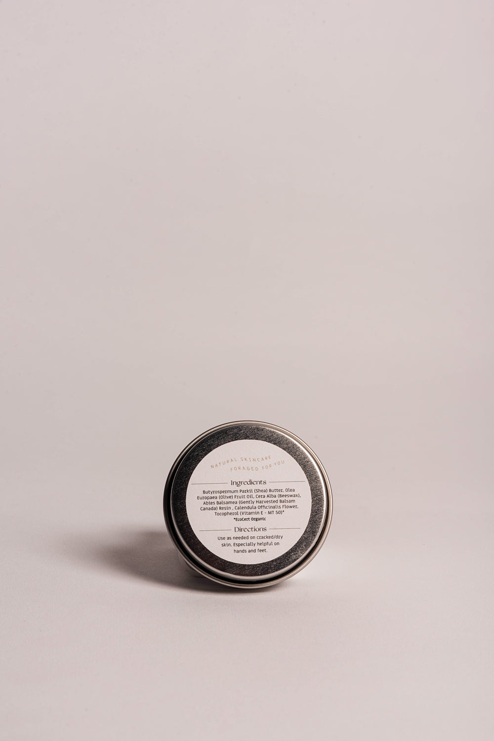 The Cedar Nook Balsam Fir Healing Salve for dry and cracked hands and feet in aluminum tin showing back label of organic ingredients with light pink background