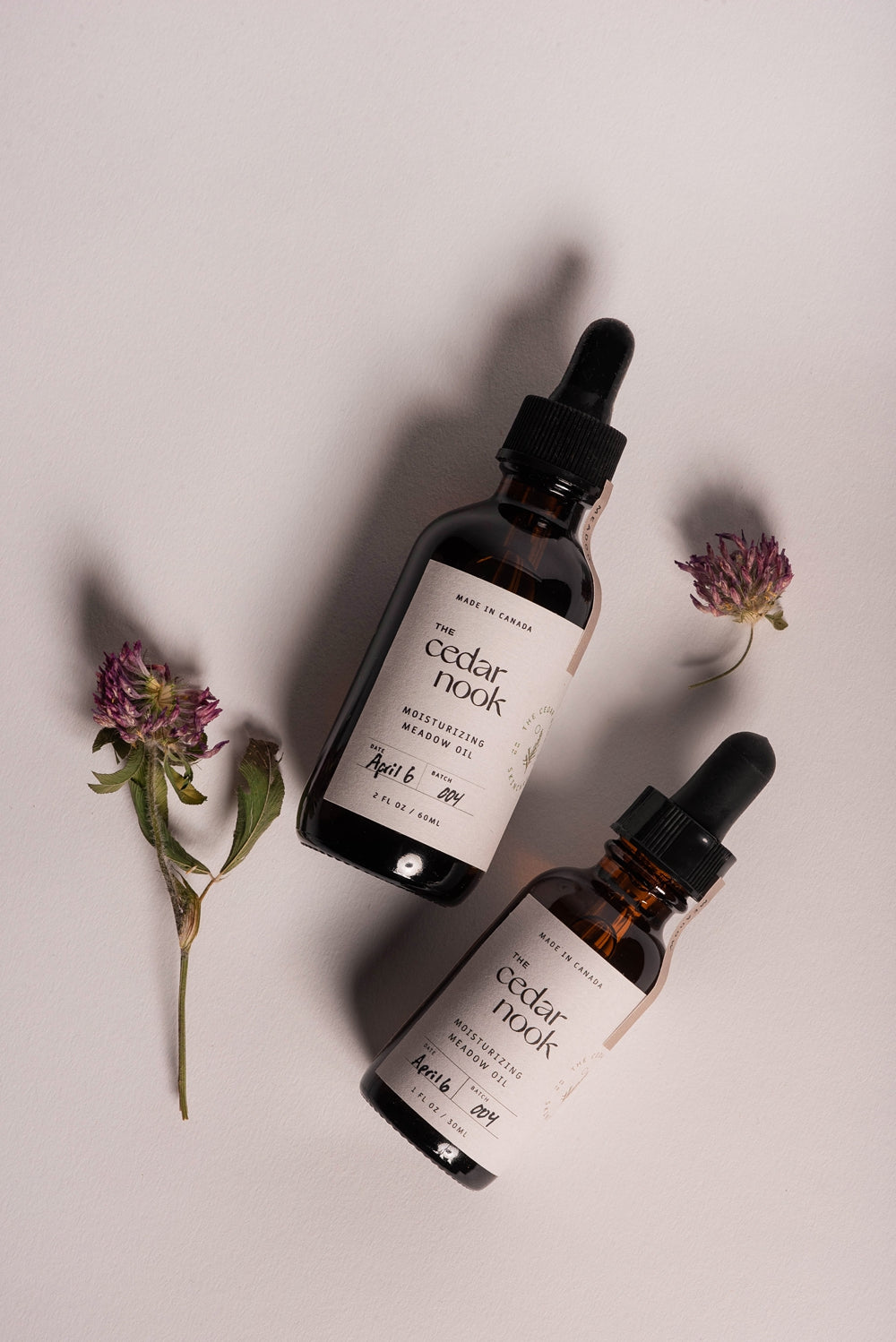 Moisturizing Meadow Oil | The Cedar Nook Face Oil both sizes 1 oz and 2 oz amber glass bottles with dropper tops showing a flat lay view surrounded by foraged red clover blossoms