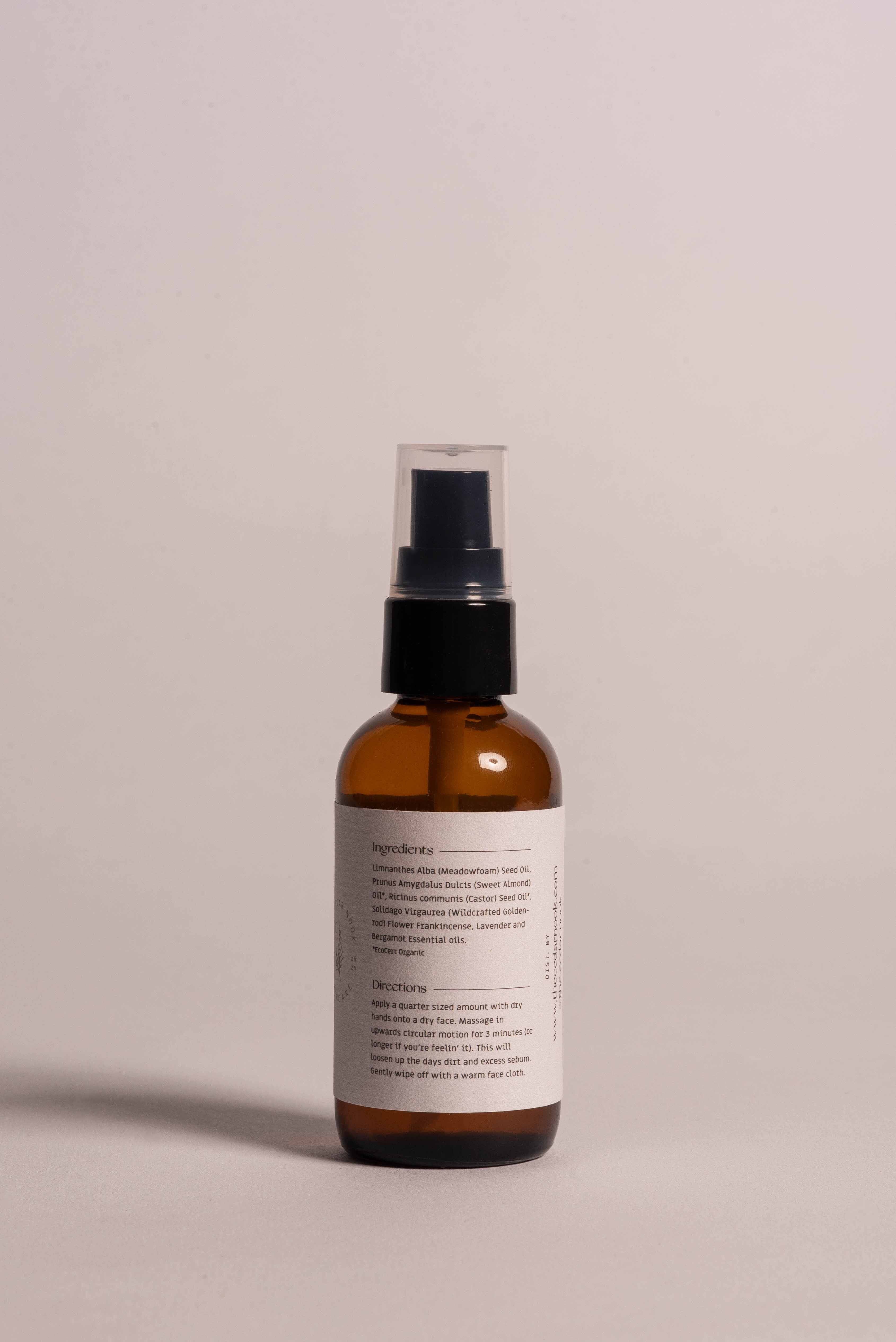 The Cedar Nook Goldenrod Oil Cleanser in 2 oz reusable amber glass bottle with airless pump top showing back label of organic ingredients and directions of use on light pink backdrop