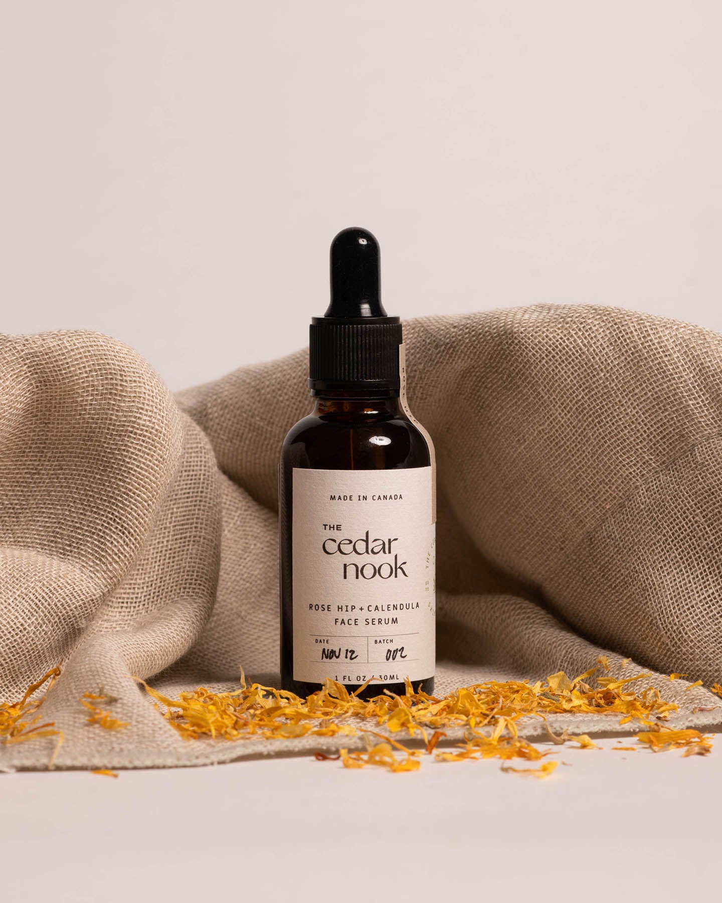 Rose Hip and Calendula Face Serum in a 1 oz amber glass bottle, standing in front of a burlap sack and surrounded by dried Calendula