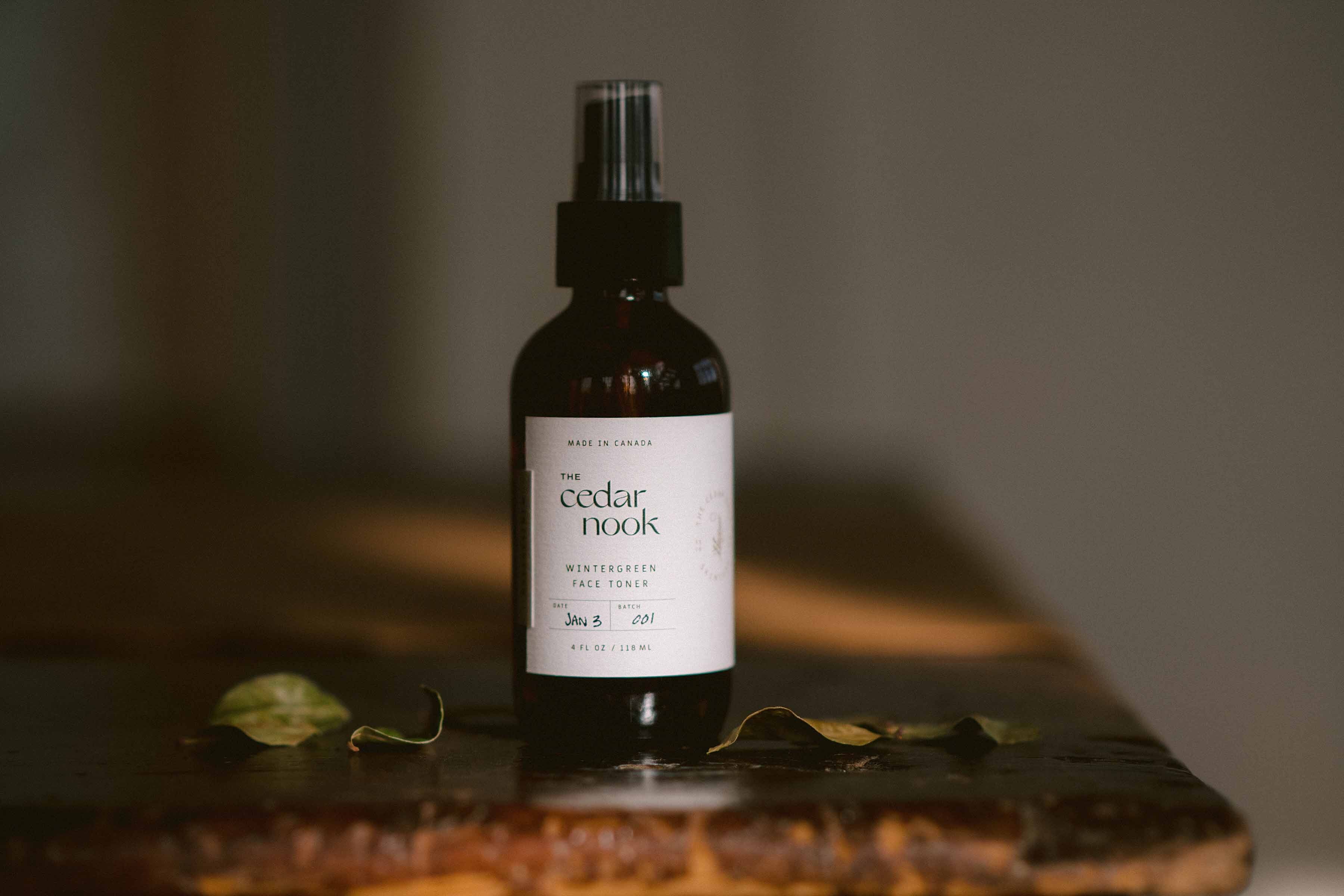 The Cedar Nook Wintergreen Face Toner in a 4 oz amber glass bottle, with fine mist spray top sitting on a wooden table surrounded by foraged wintergreen leaves 