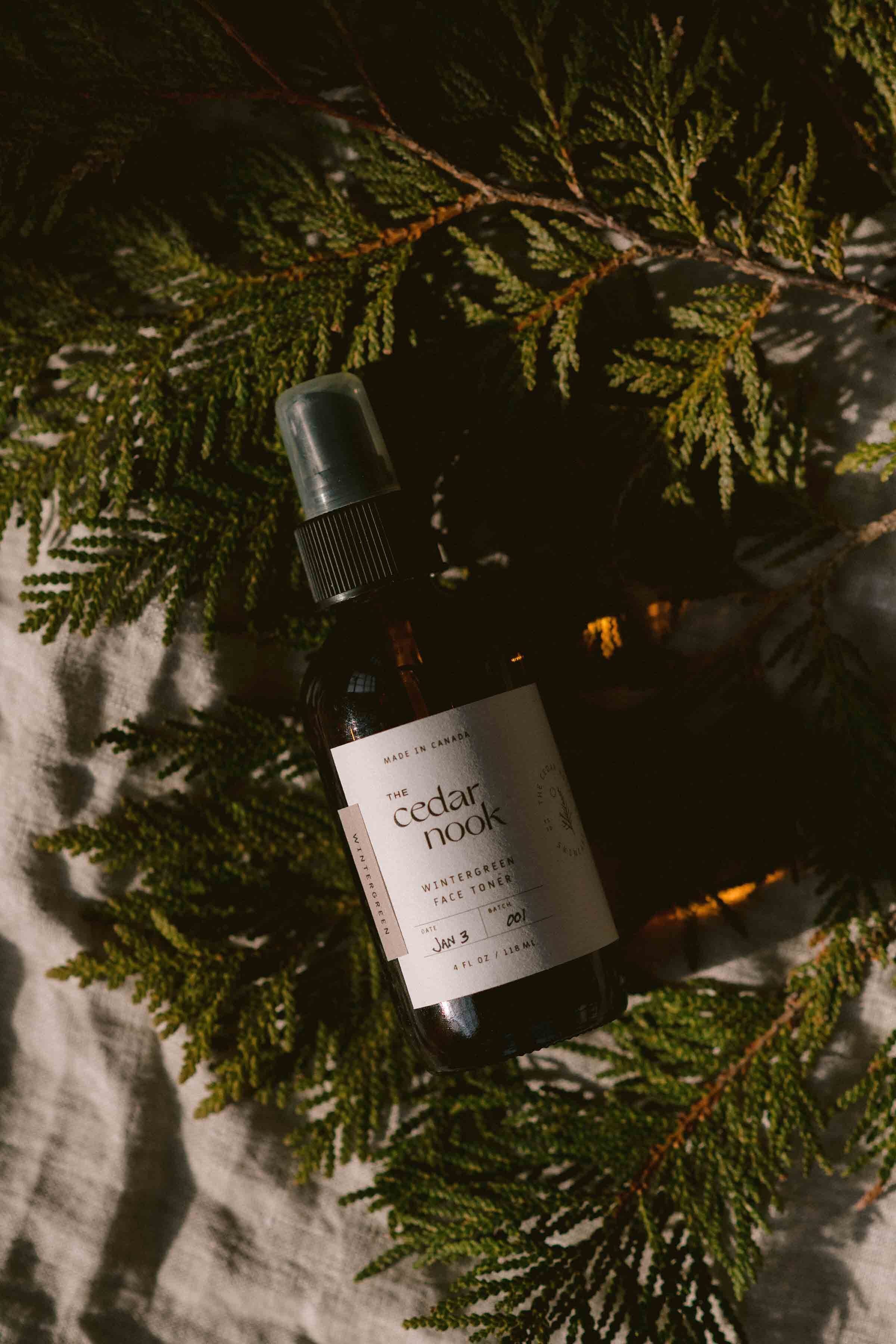 The Cedar Nook Wintergreen Face Toner in a 4 oz amber glass bottle with fine mist spray top, laying flat on a bed of fresh cedar boughs and grey linens