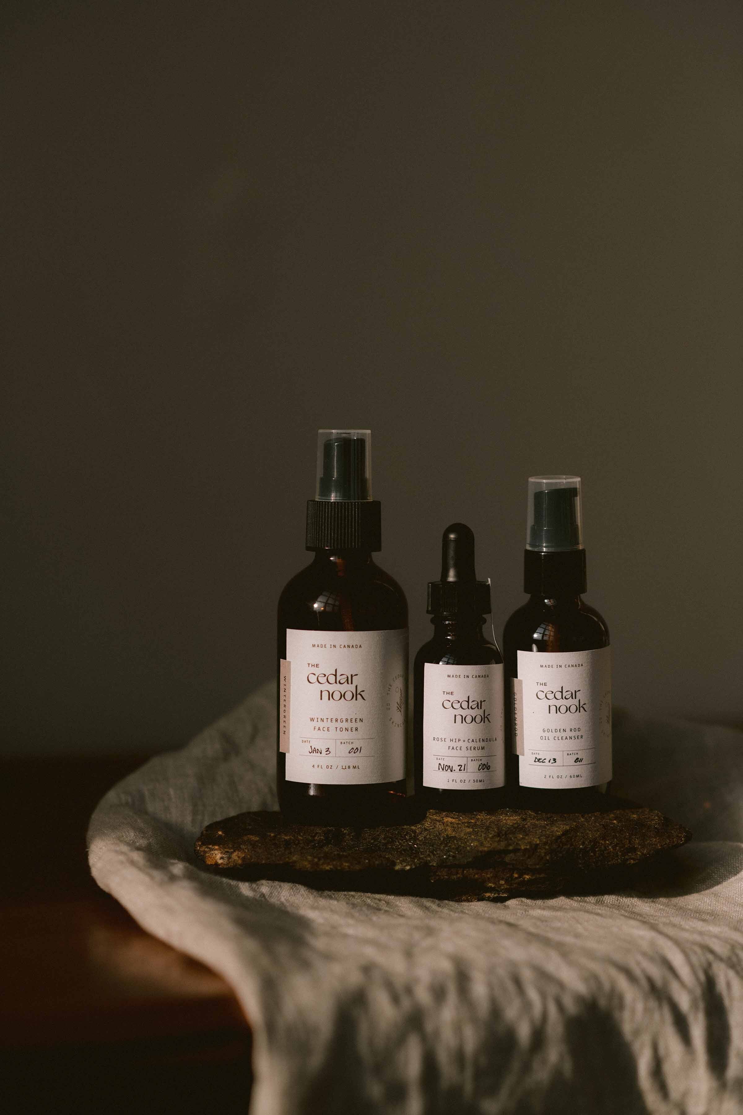 The Cedar Nook Wintergreen Face Toner with Rose Hip and Calendula Face Serum and Goldenrod Oil Cleanser. The three bottles are sitting aesthetically on a rock on a wooden table with some grey linen.