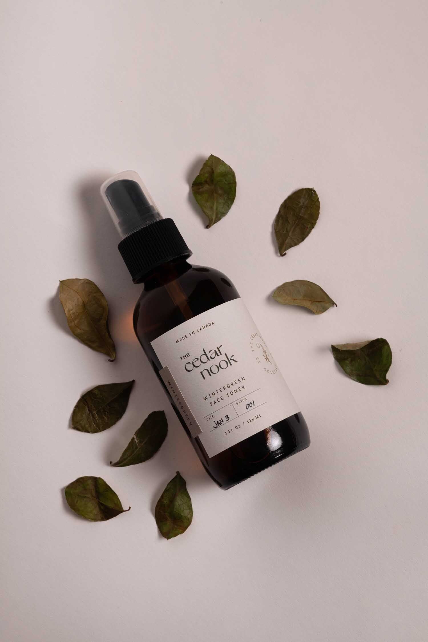 A flat lay shot of The Cedar Nook Wintergreen Face Toner in a 4 oz amber glass bottle with spray top, surrounded by foraged wintergreen leaves on a white background